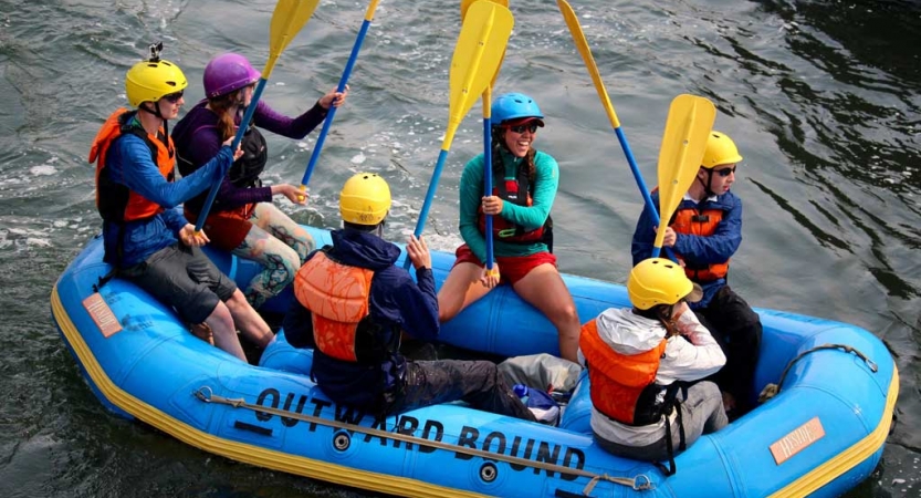 a group of outward bound students wearing helmets and life jackets sit in a raft and raise their paddles into the air